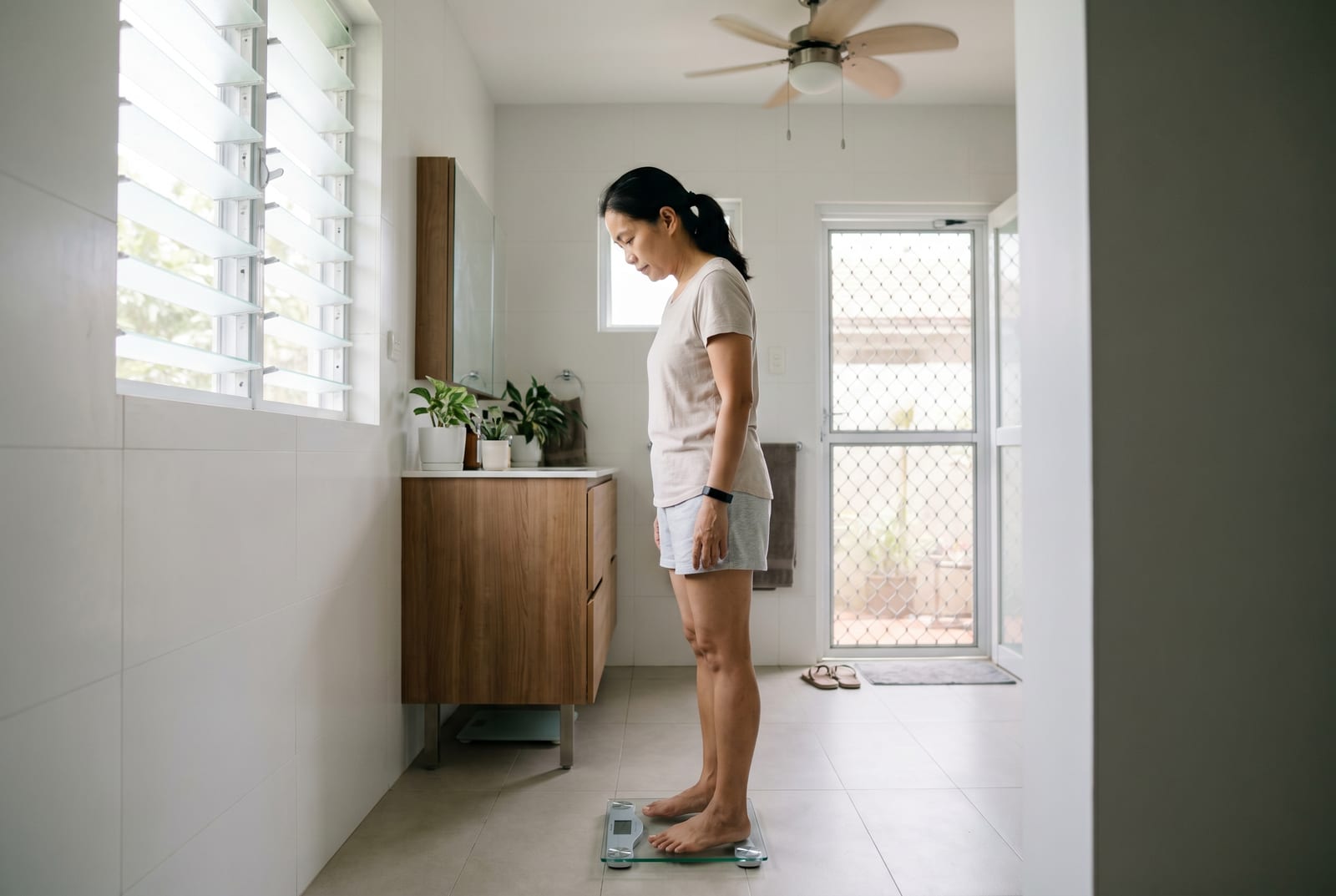 Babae sa malambot na ilaw sa banyo na bumababa sa isang smart scale, tinitingnan ang readout - AI-generated