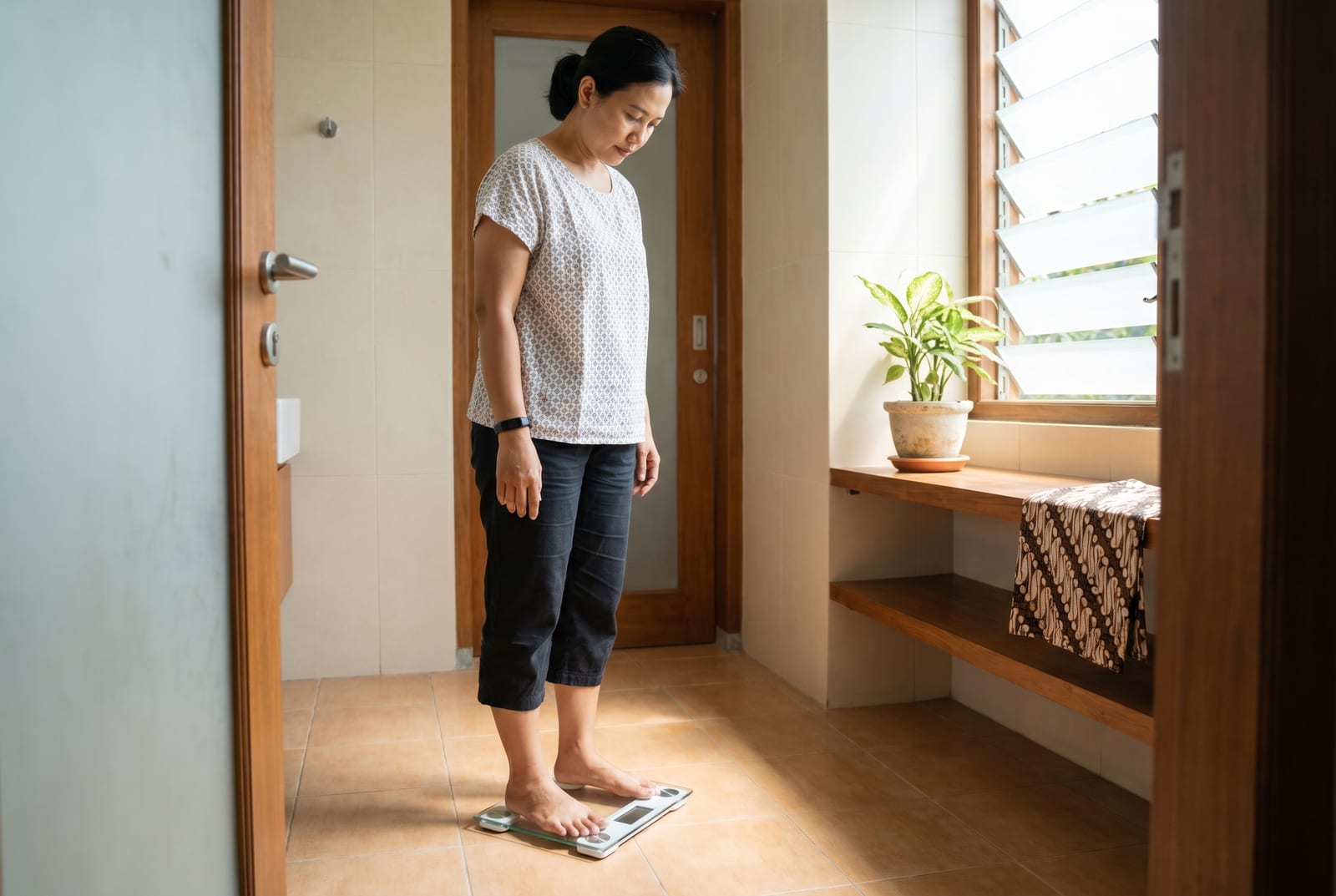 Wanita di kamar mandi dengan pencahayaan lembut, baru saja turun dari timbangan pintar, melirik angka yang tertera - Dibuat oleh AI